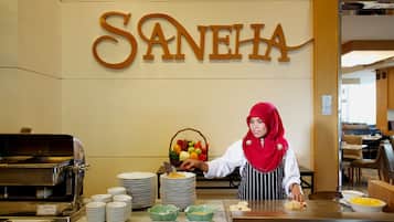 Montaje de bufé con un letrero que dice "SANEHA" en la parte superior, una barra de bufé con platos y recipientes, y un miembro del personal sirviendo comida.