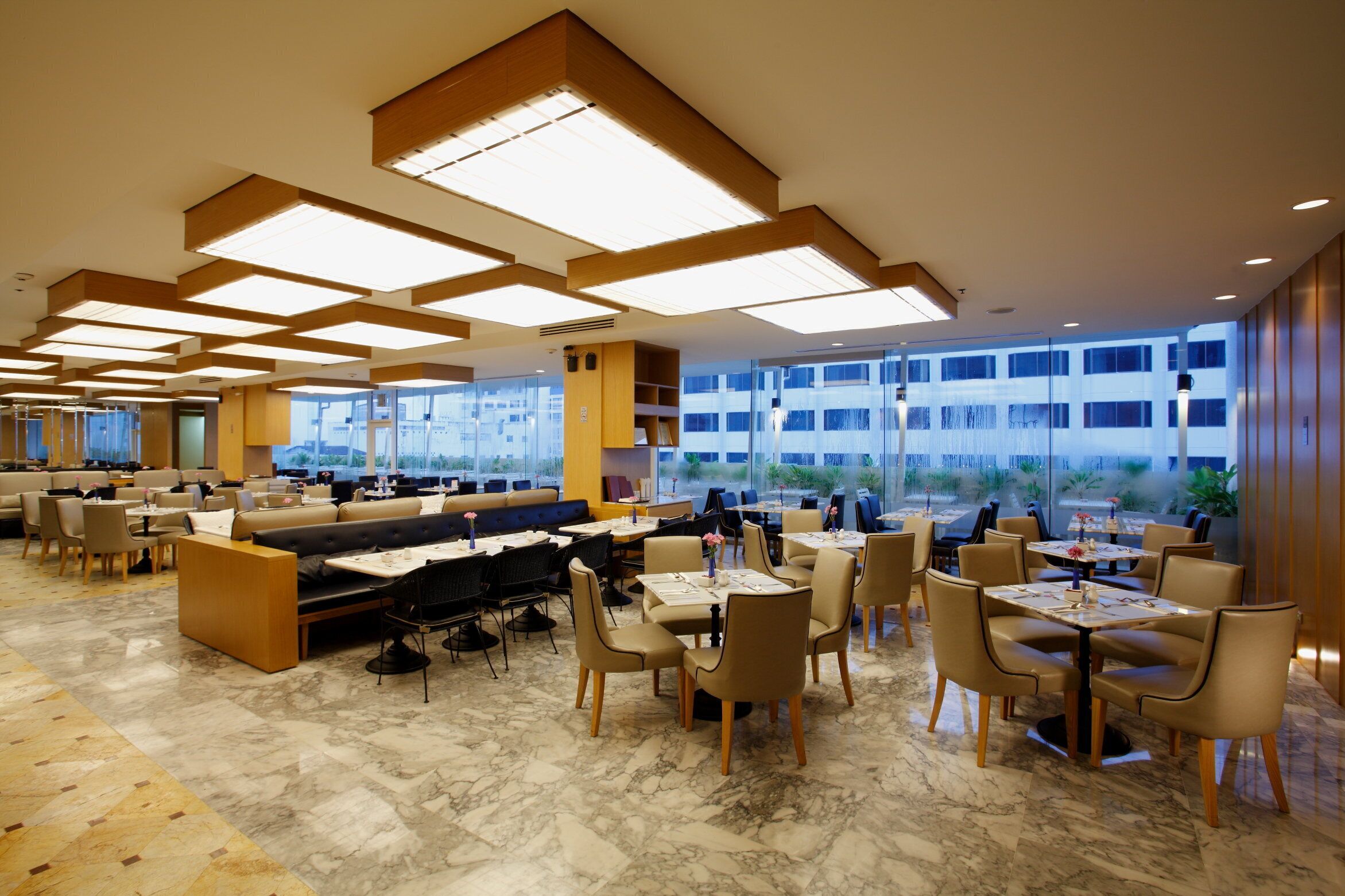 A modern restaurant with marble flooring, wooden furniture, and large windows offering a view of the city.
