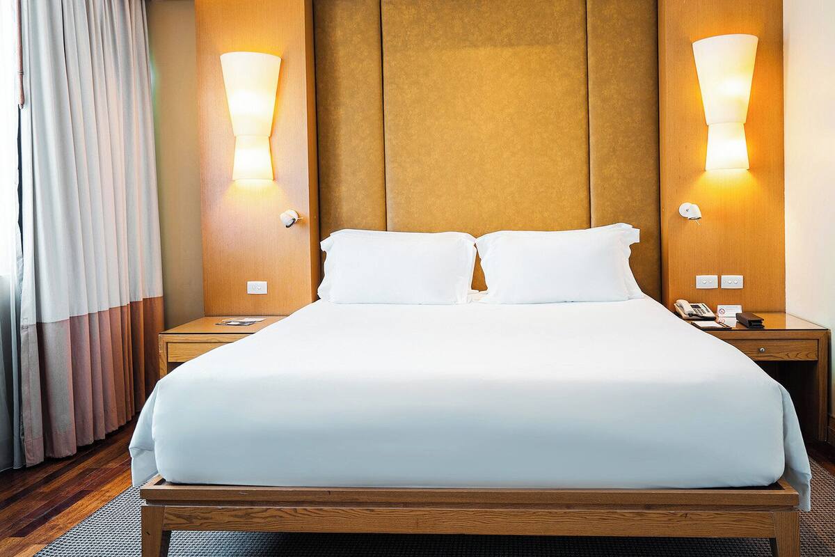 A hotel room with a large bed, wooden headboard, two bedside lamps, a nightstand with a phone and a notepad, and a window with curtains.