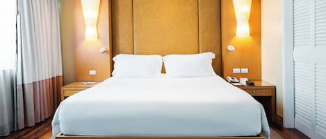 A hotel room with a large bed, wooden headboard, two bedside lamps, a nightstand with a phone and a notepad, and a window with curtains.