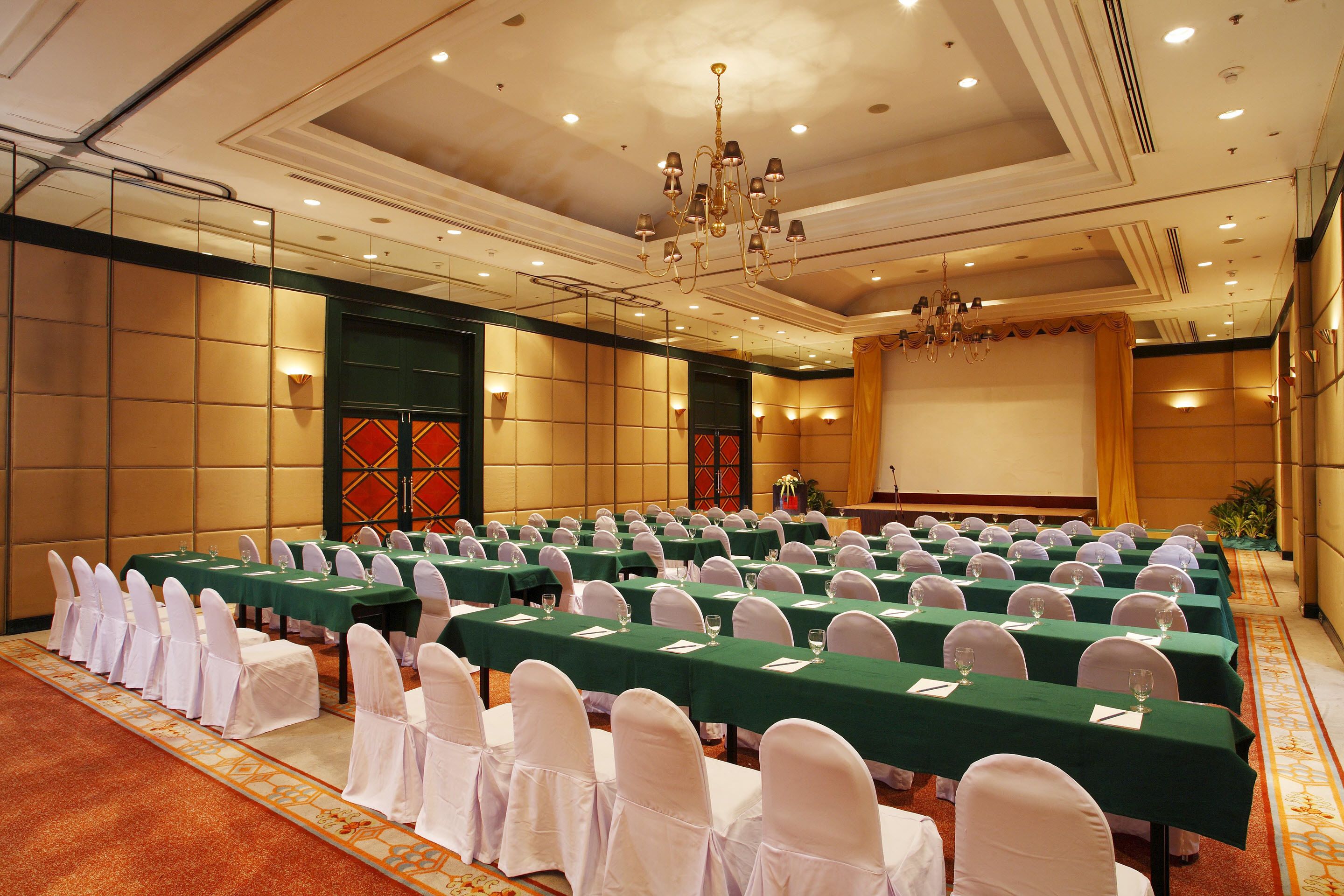 A conference room with rows of tables and chairs, a chandelier, and a large screen.