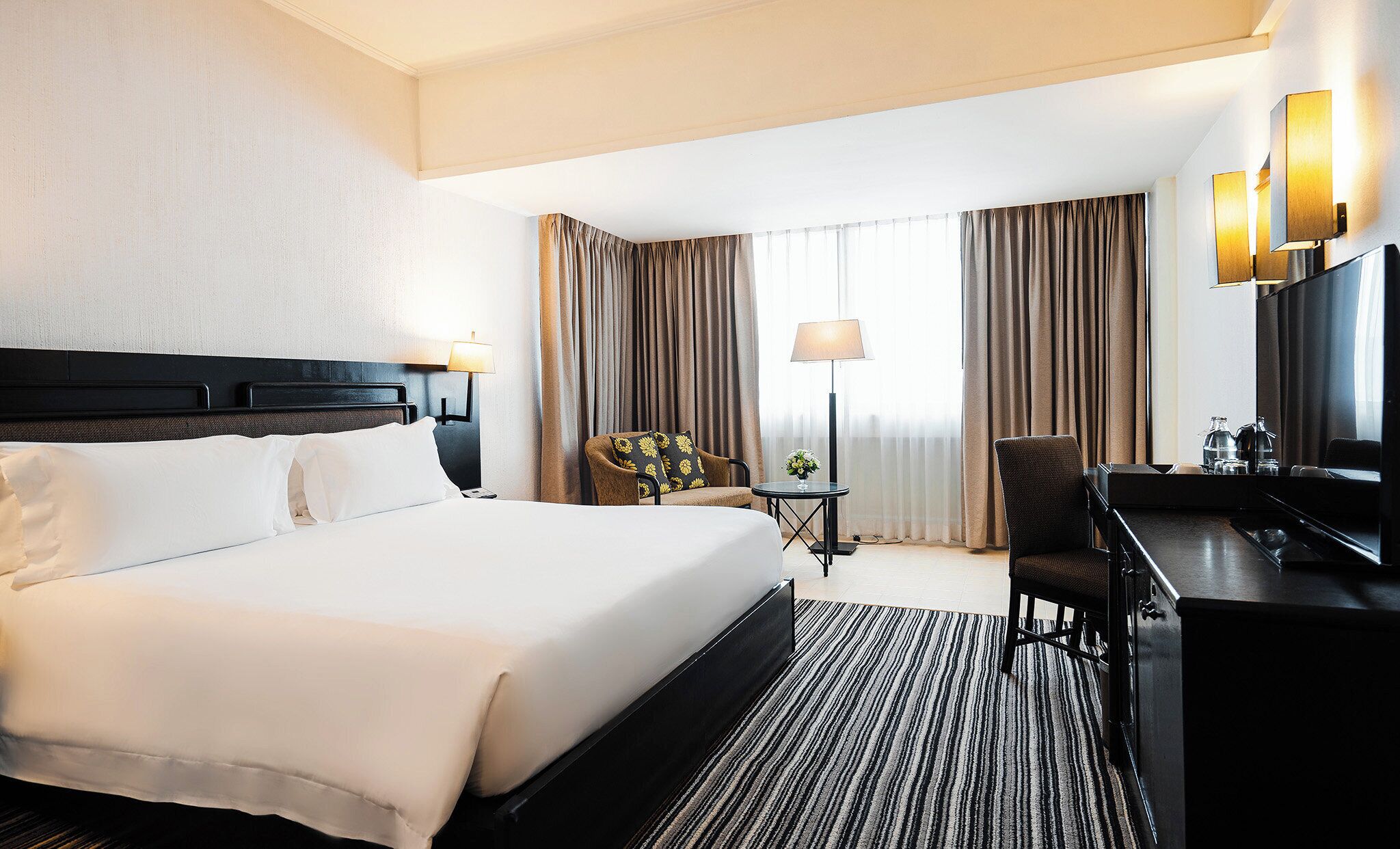 A hotel room with a large bed, a desk, a chair, a lamp, and curtains.
