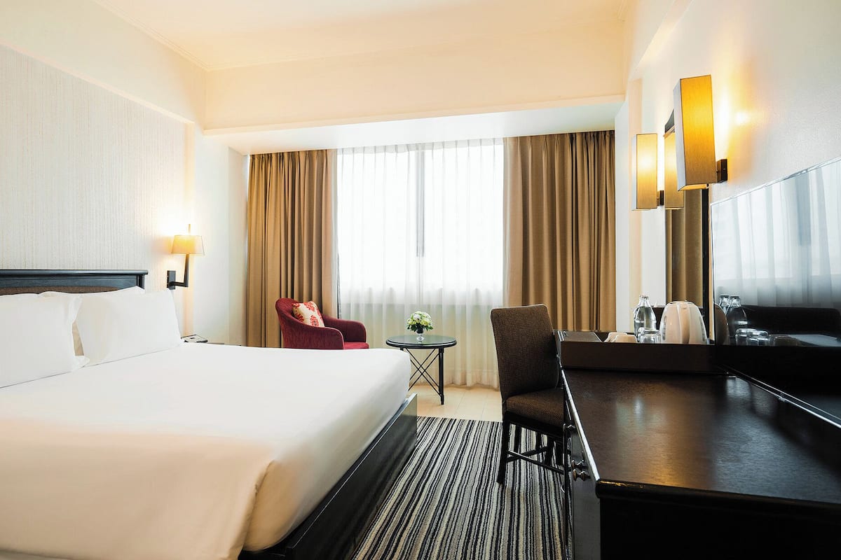 A hotel room with a large bed, a desk, a chair, a small table with a vase of flowers, and a large mirror.