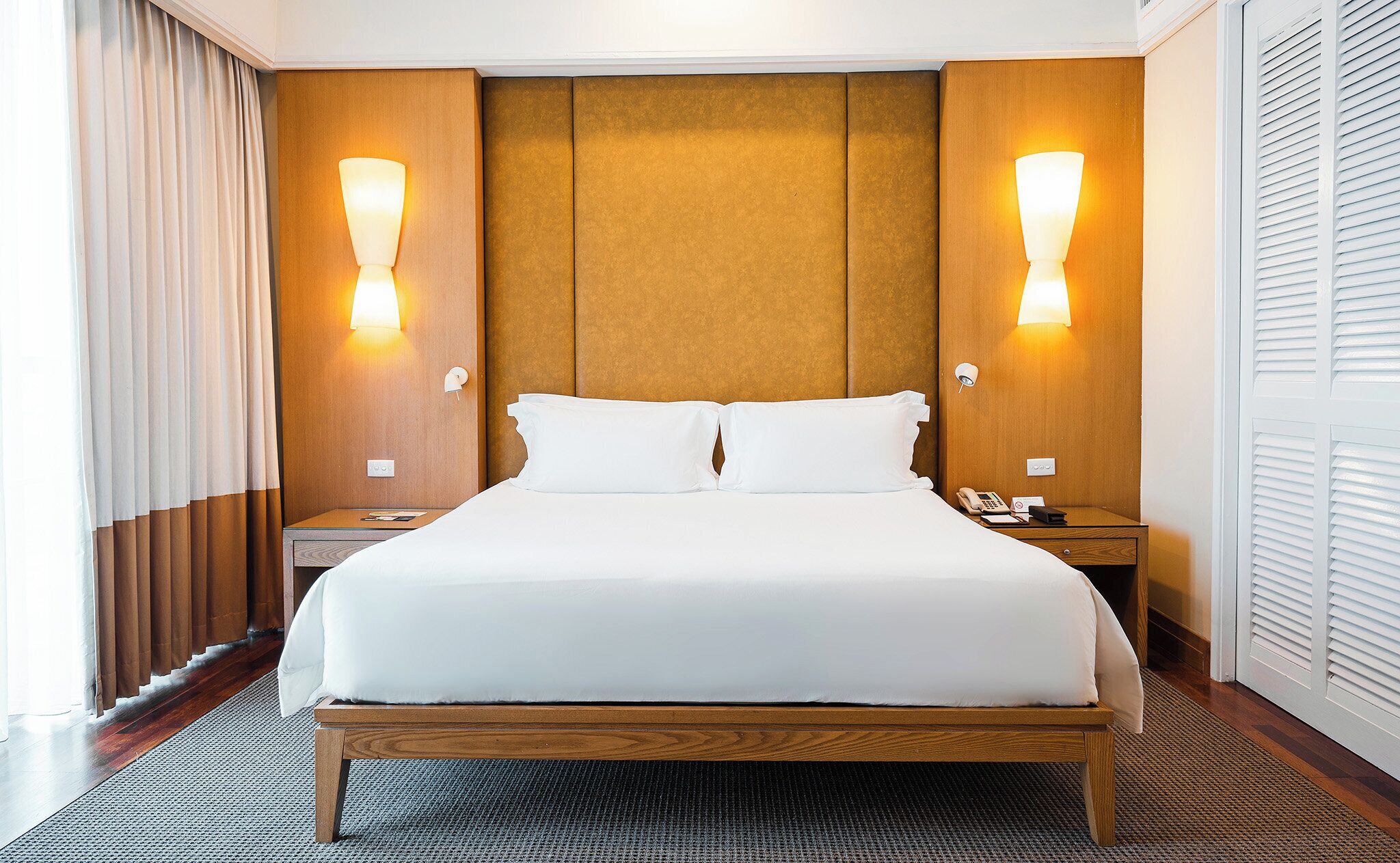 Ein Hotelzimmer mit einem großen Bett, einem Holzkopfteil, zwei Nachttischlampen, einem Nachttisch mit Telefon und Notizblock sowie einem Fenster mit Vorhängen.