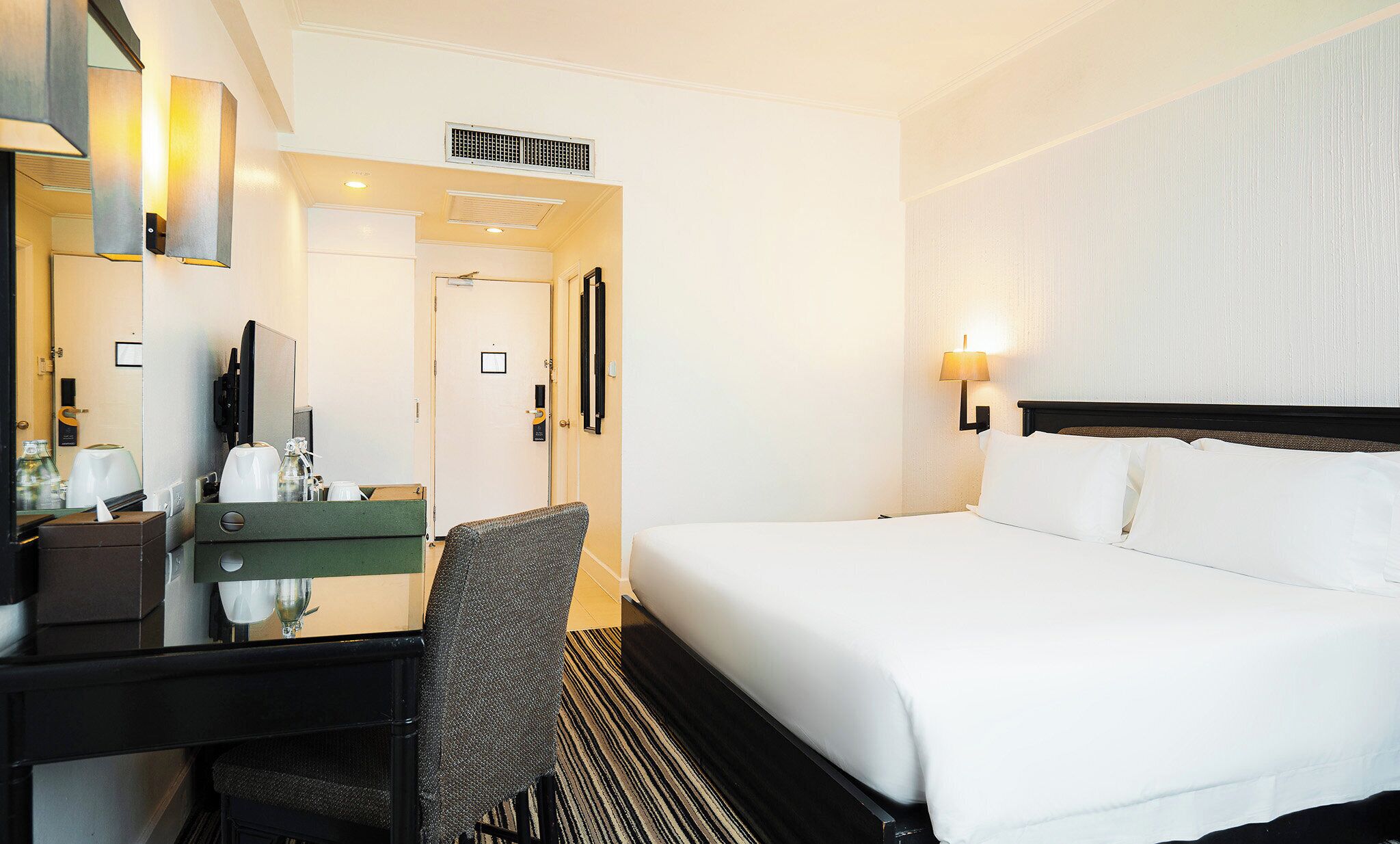 A hotel room with a bed, a desk with a chair, a mirror, and a wall-mounted lamp.