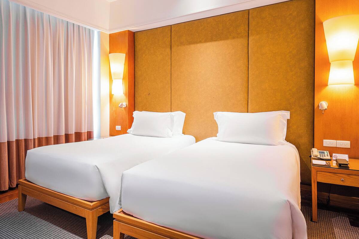 A hotel room with two single beds, a wooden headboard, a bedside table with a phone and a notepad, and a window with curtains.