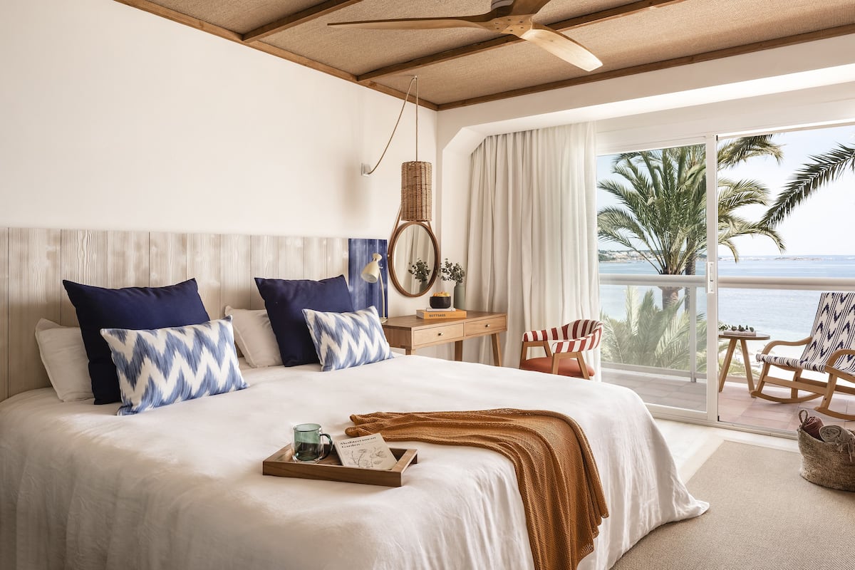 A bedroom with a large bed, a bedside table, a chair, and a view of the ocean.