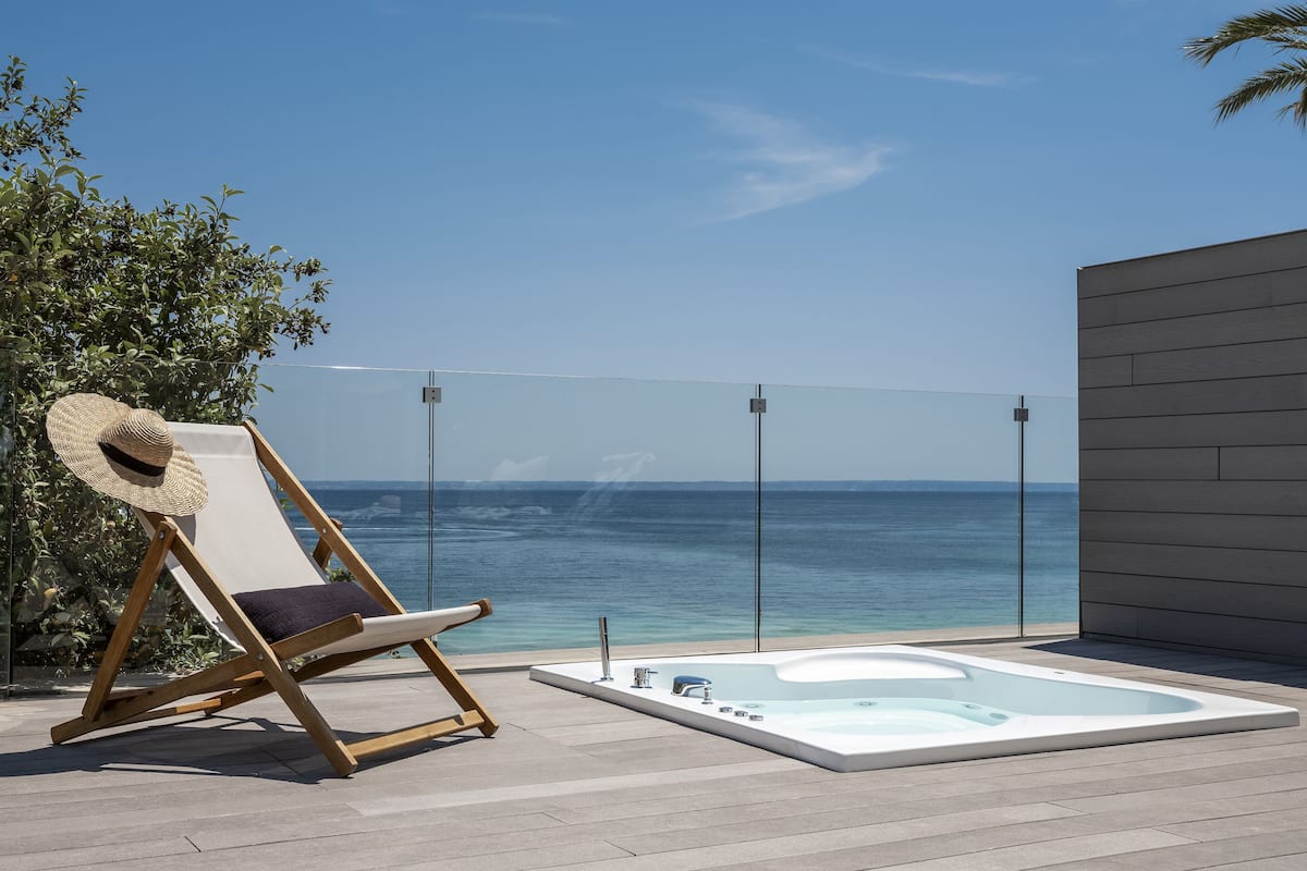 A wooden lounge chair with a straw hat, a hot tub, and a clear view of the ocean.