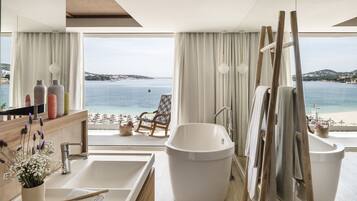 A modern bathroom with a large bathtub, a sink, and a shower area. It features a large window with a view of the sea and a beach.