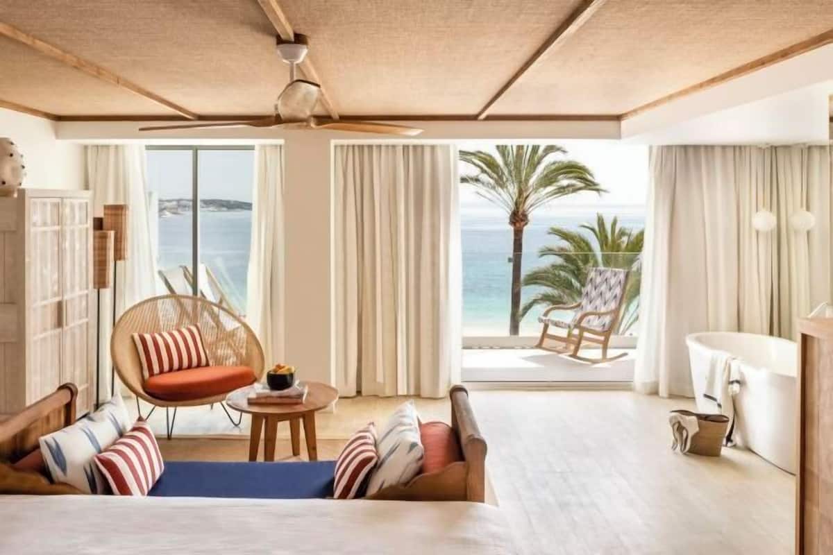 A spacious room with a large bed, a sitting area with a chair and a table, a bathtub, and a view of the ocean.