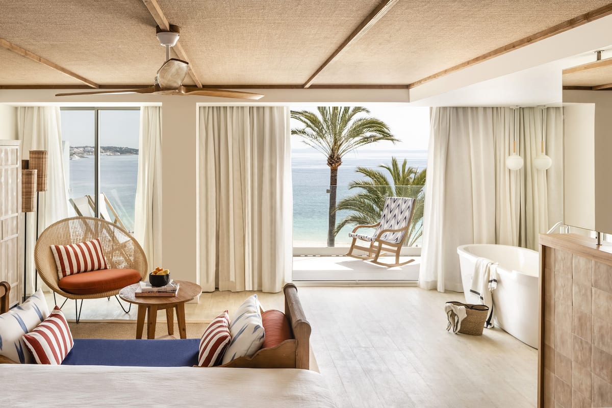 A spacious room with a large bed, a seating area with a wicker chair, a small table, and a view of the ocean.