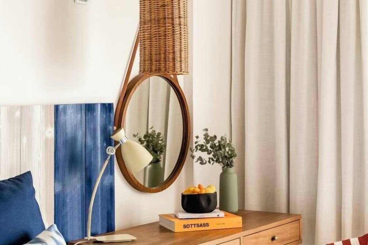 A neatly arranged hotel room with a wooden nightstand, a mirror, a lamp, a book titled 'SOTTSASS,' and a vase with flowers.