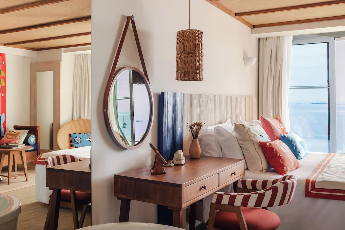 A hotel room with a bed, a desk, a chair, a bathroom, and a view of the sea.