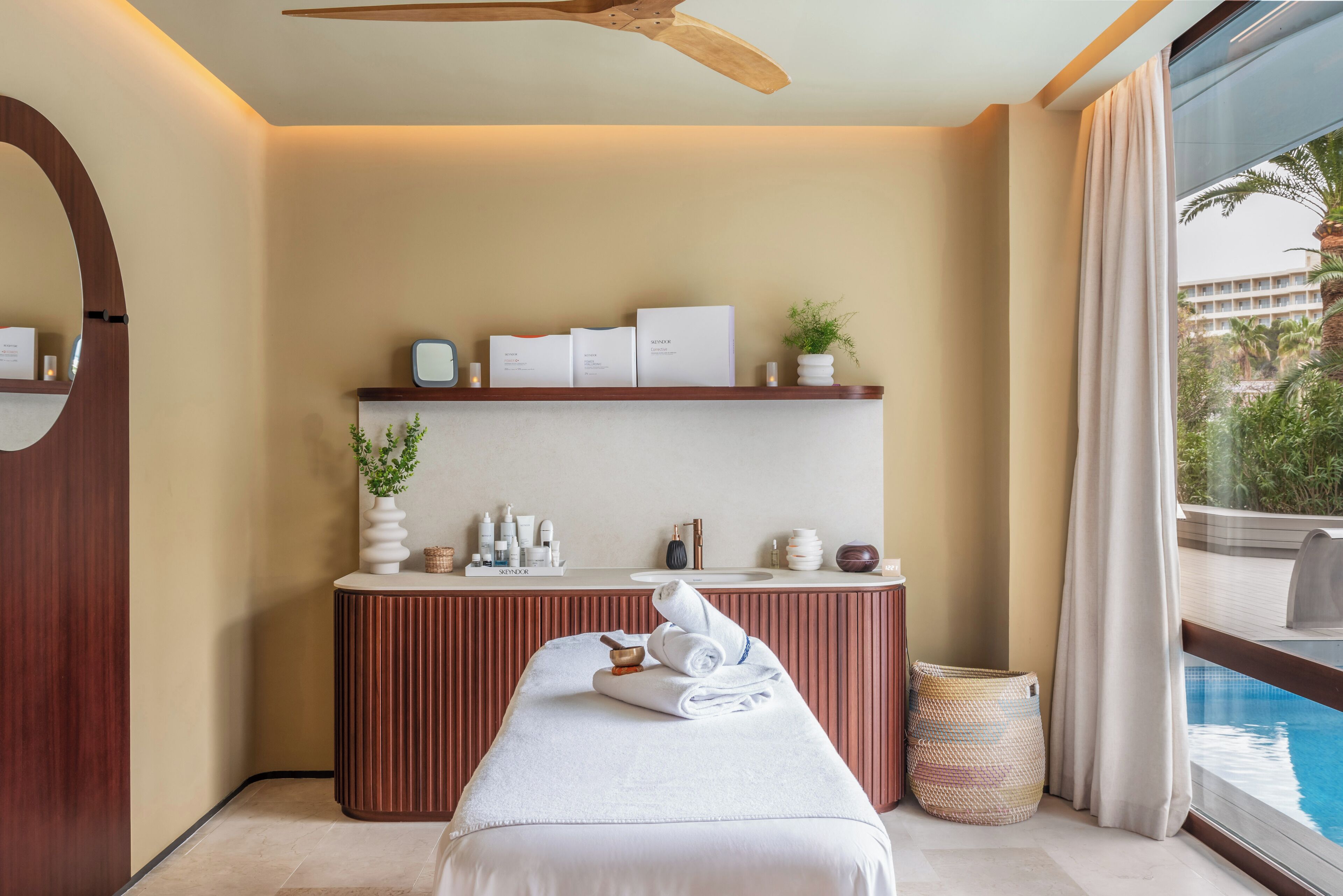 A hotel room with a bed, a vanity with toiletries, a shelf with books, a ceiling fan, and a view of a swimming pool.