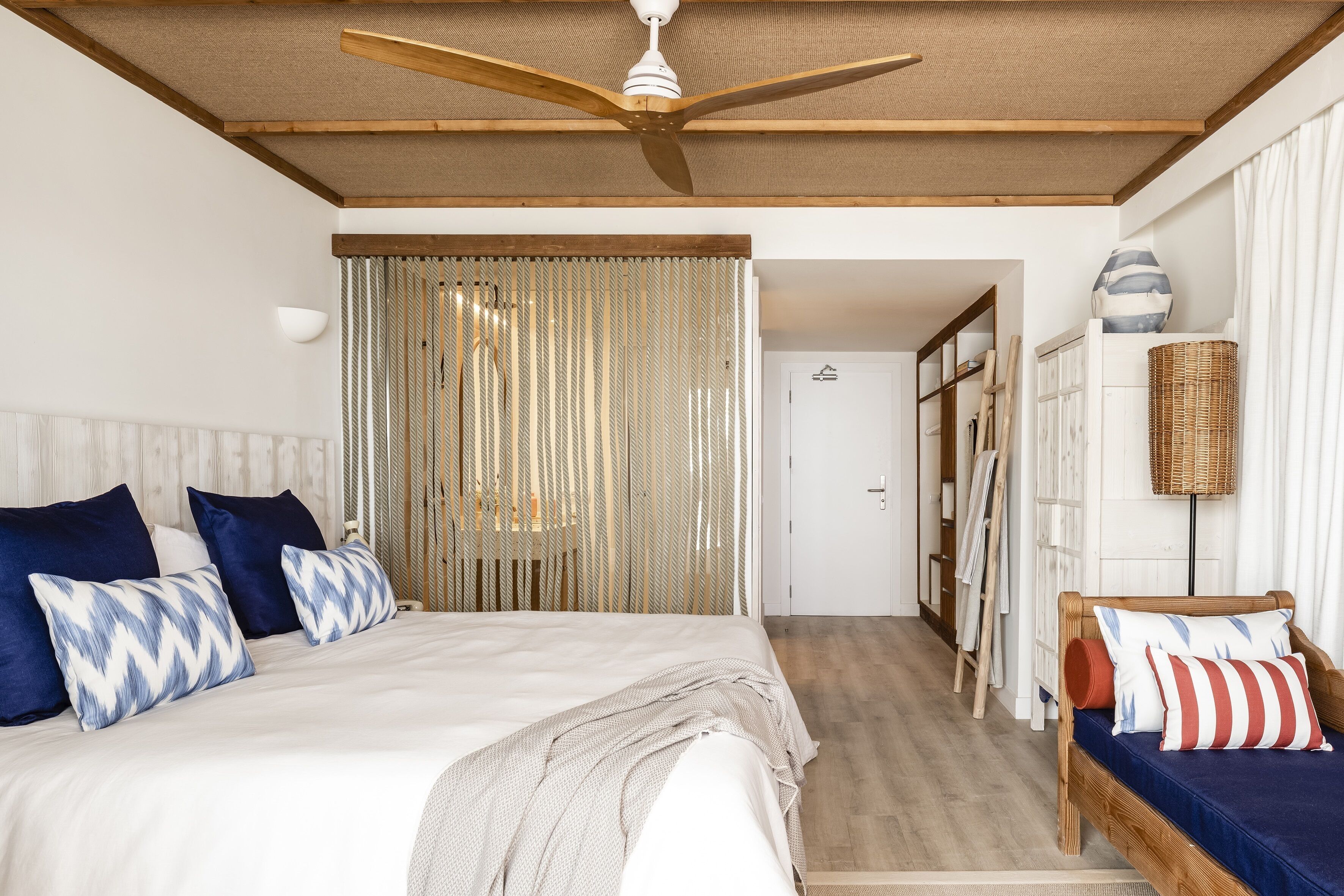 A bedroom with a large bed, a wooden ceiling fan, a woven curtain, and a small seating area with cushions.