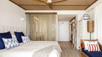 A bedroom with a large bed, a wooden ceiling fan, a woven curtain, and a small seating area with cushions.
