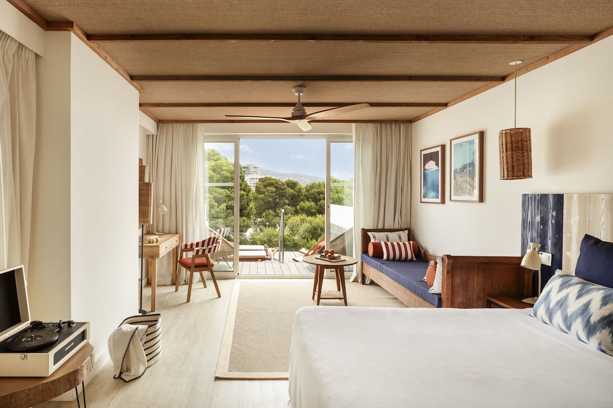 A hotel room with a bed, a desk, a chair, a sofa, and a balcony with a view of greenery and buildings.