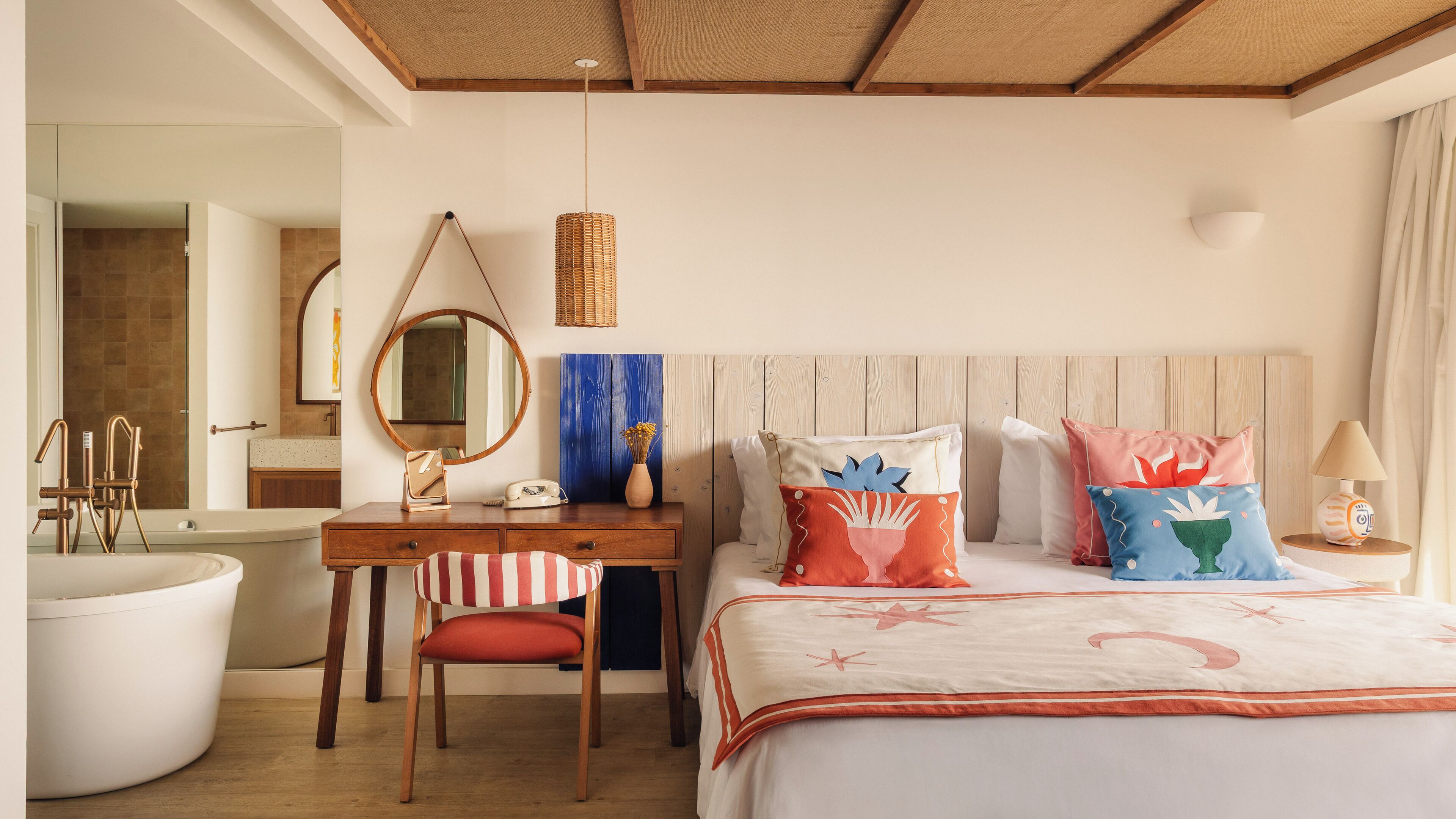 A hotel room with a wooden ceiling, a large bed with colorful pillows, a desk with a chair, a round mirror, a wicker lamp, and a bathroom with a bathtub and a toilet.