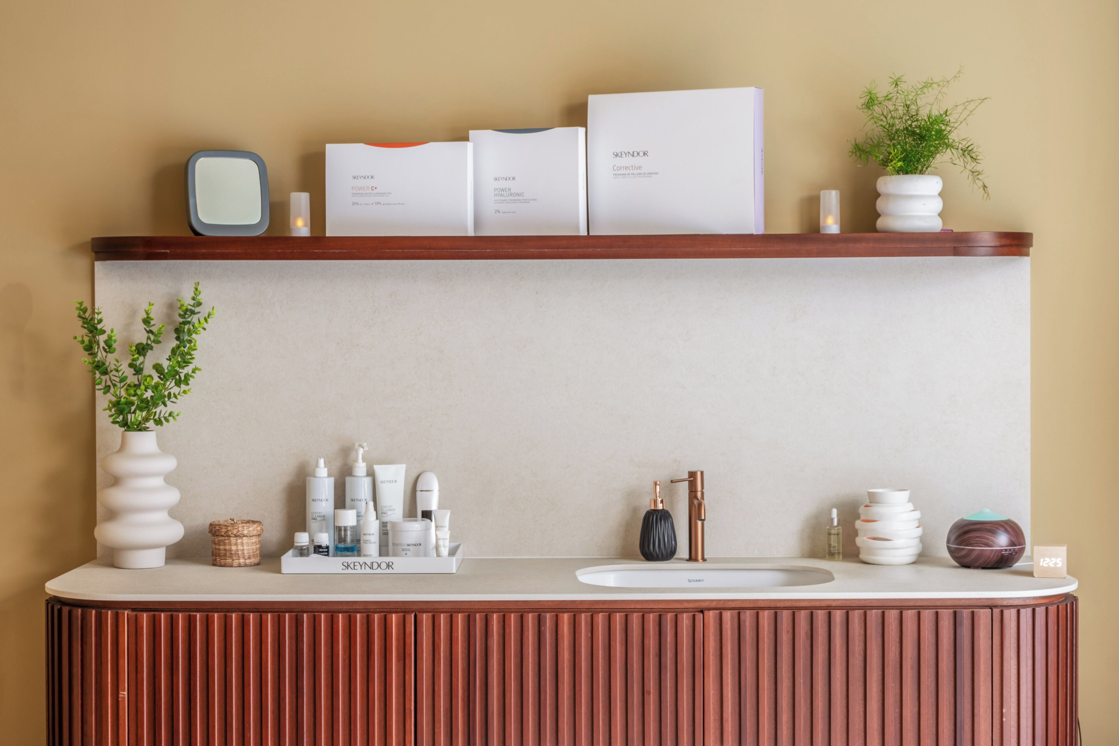 A modern bathroom vanity with a wooden base, a white countertop, and a shelf above holding skincare products and a potted plant.