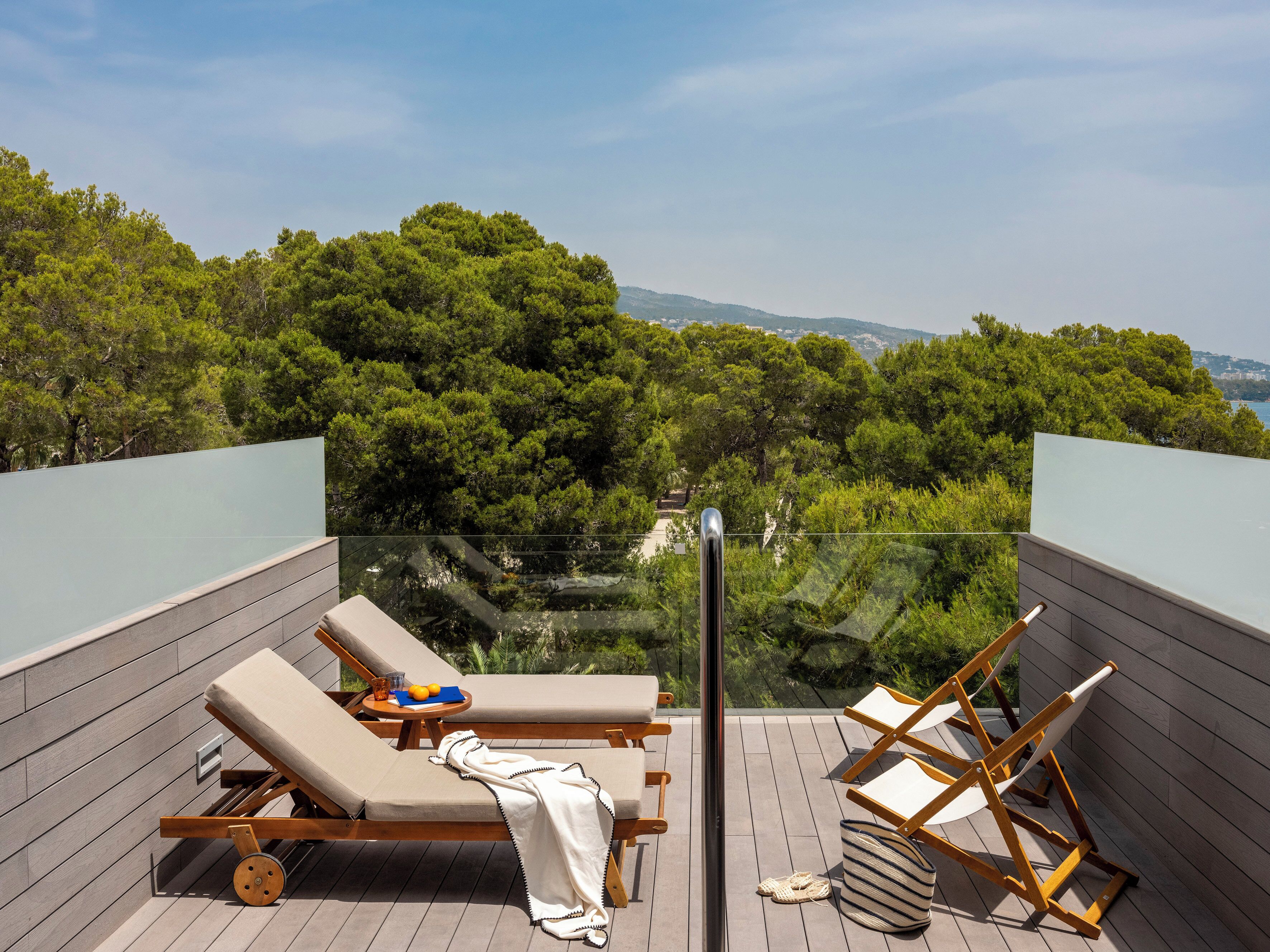 A balcony with two lounge chairs, a small table with a drink and fruit, and a view of a forested area.