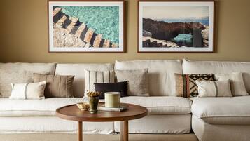 A living room with a beige sofa, two framed coastal landscape paintings, a round wooden coffee table with a vase and books, and a textured rug.