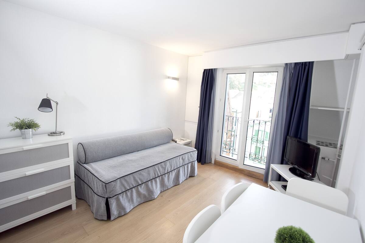A modern, well-lit room with a grey sofa, a small white table, a TV on a stand, and a balcony with a view.