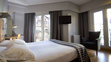 Habitación de hotel con una cama grande, un televisor de pantalla plana montado en la pared, una silla y vistas a la ciudad a través de las ventanas.