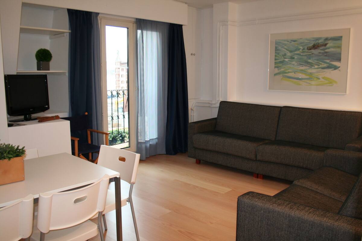 A modern living room with a grey sofa, a dining table with chairs, a TV on a stand, and a balcony with blue curtains.