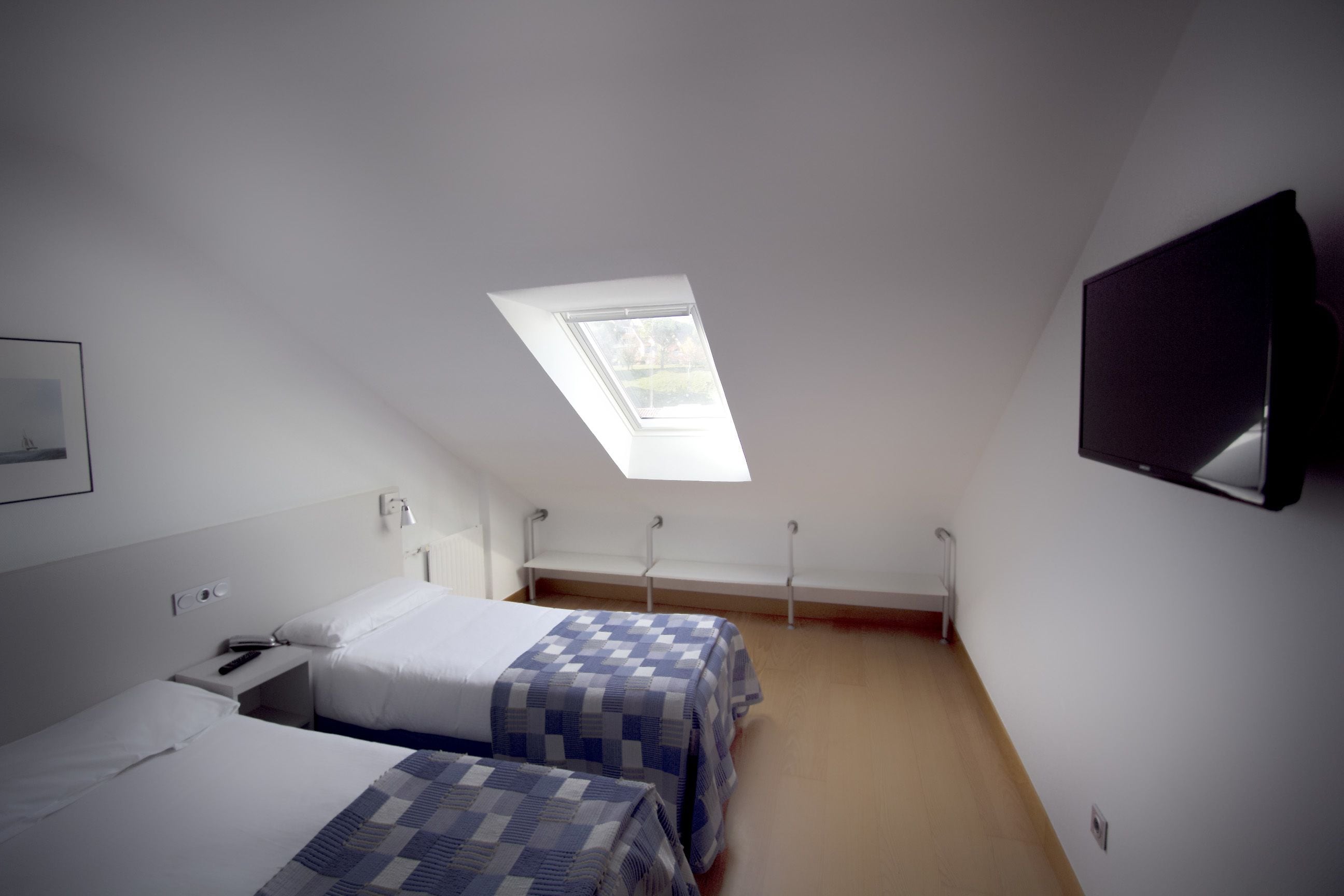 Un dormitorio con dos camas, un tragaluz, un televisor montado en la pared y una mesita de noche con lámpara.