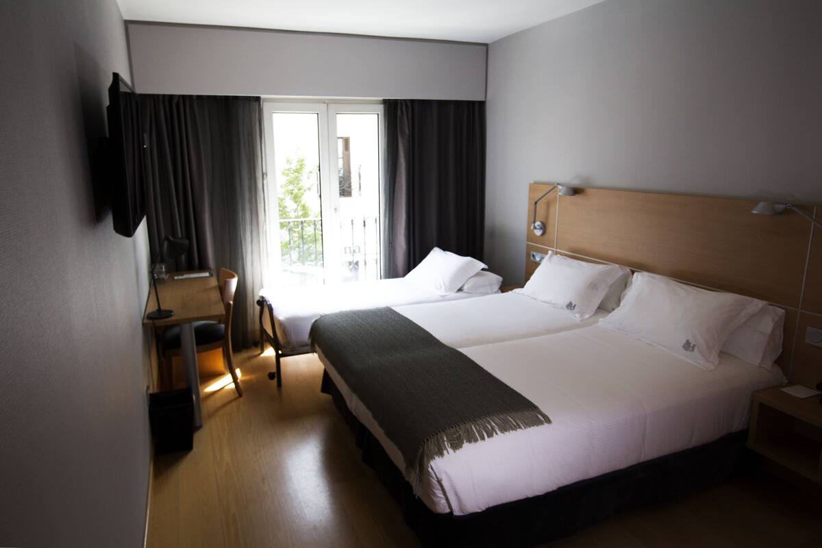 A hotel room with a large bed, a desk with a chair, a TV mounted on the wall, and a window with curtains.
