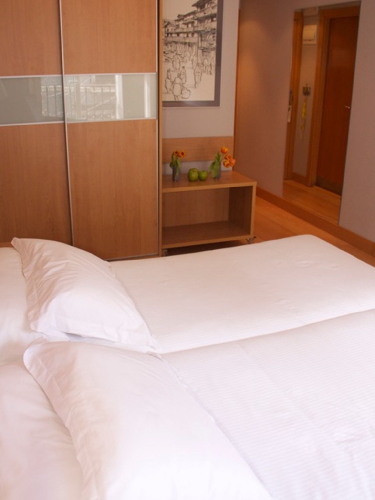 A bedroom with a bed, wooden wardrobe, a small wooden table with decorative items, and a framed picture on the wall.