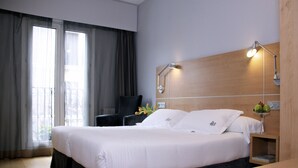 A hotel room with a large bed, a desk, a chair, a nightstand, a lamp, and a view of the outside through the window. - Hotel Jauregui (Hondarribia)