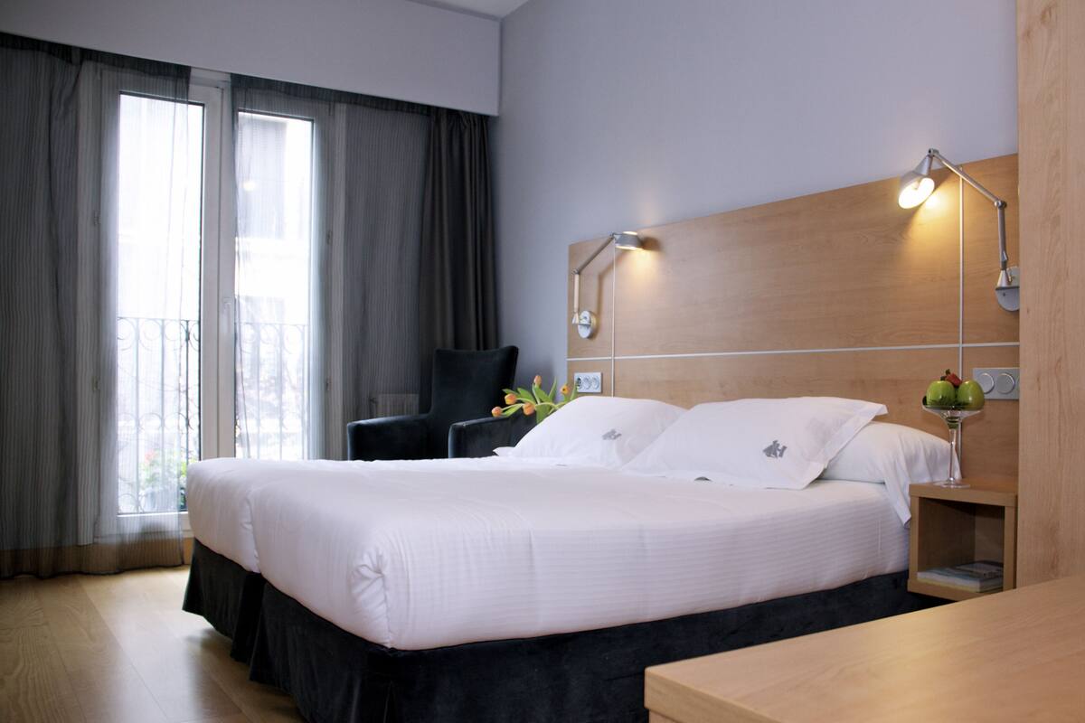 A hotel room with a large bed, a desk, a chair, a nightstand with a lamp, and a window with curtains.