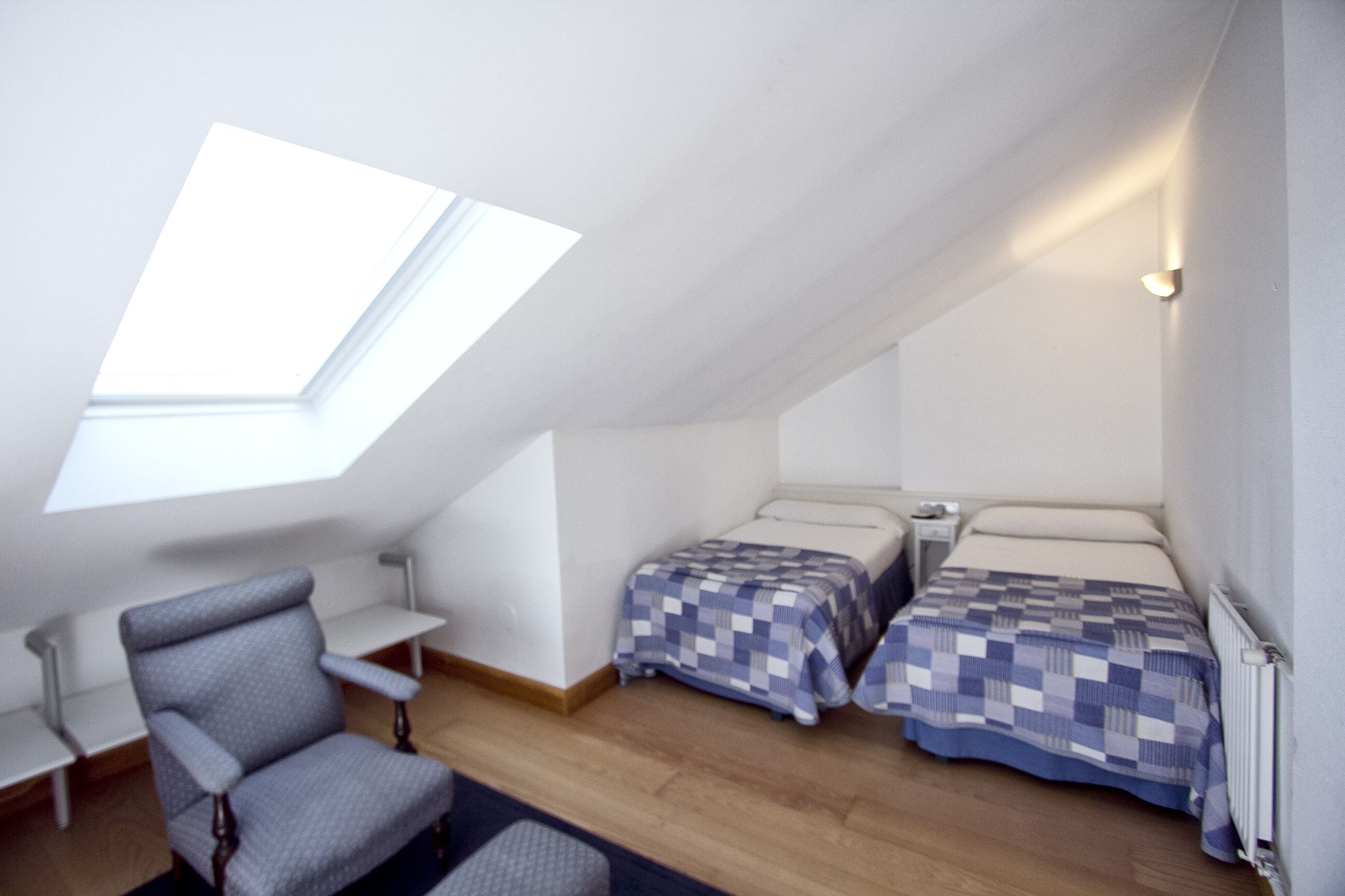 Una habitación pequeña y acogedora con techo inclinado, un tragaluz, dos camas con ropa de cama a cuadros, una silla, una mesita y una lámpara de noche.