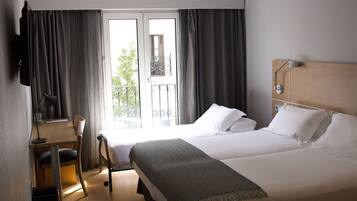 Habitación de hotel con cama, escritorio, silla, televisión y balcón con cortinas.