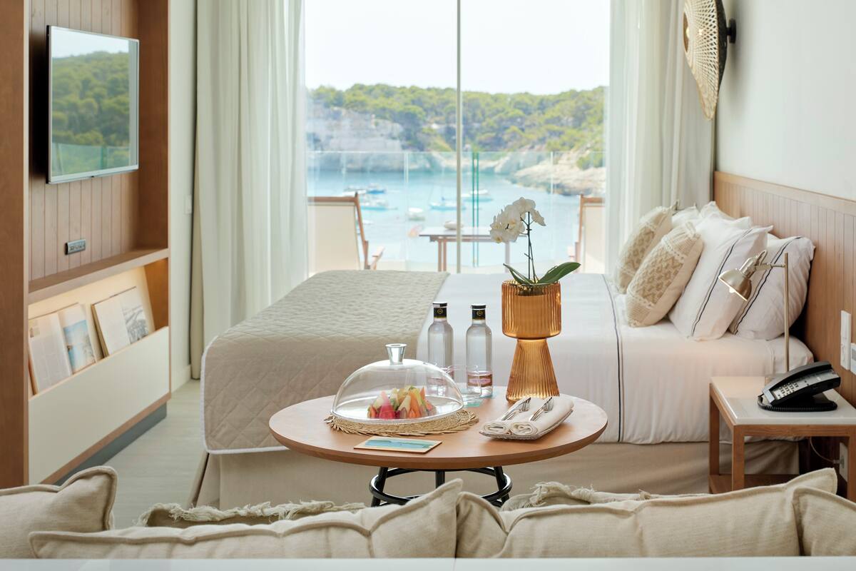 Ein Hotelzimmer mit Bett, einem runden Tisch mit Glaskuppen-Dekoration, einer Couch mit Kissen, einem an der Wand befestigten Fernseher und Meerblick durch große Fenster.