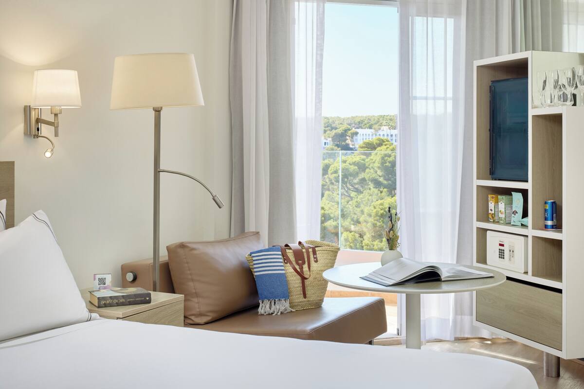 Ein modernes Hotelzimmer mit einem Bett, einem Nachttisch, einem Schreibtisch zum Lesen, einem Bücherregal und Blick auf Grünfläche.