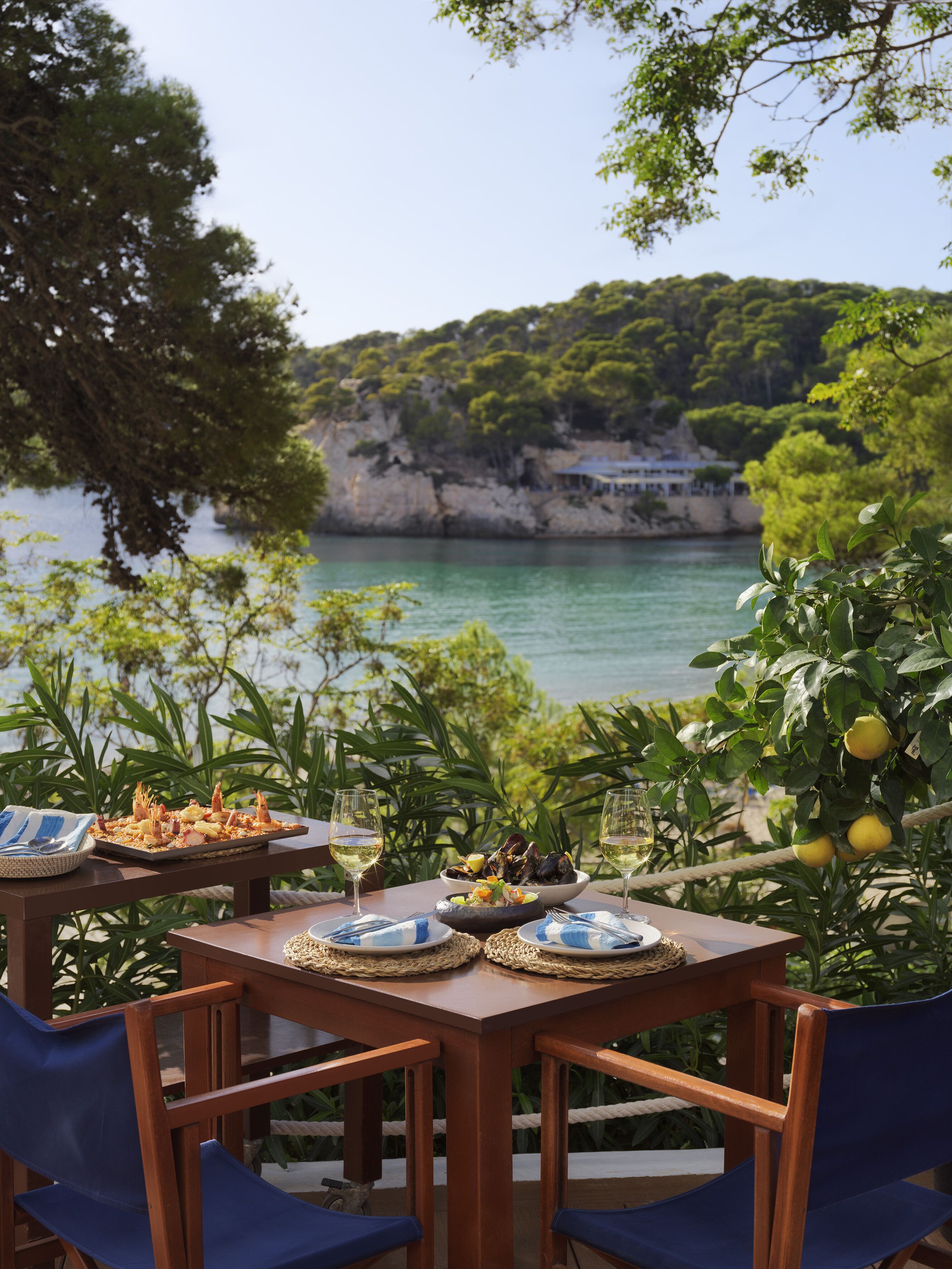 Mesa posta com pratos de frutos do mar, copos de vinho e vista para paisagem costeira com falésias e vegetação.