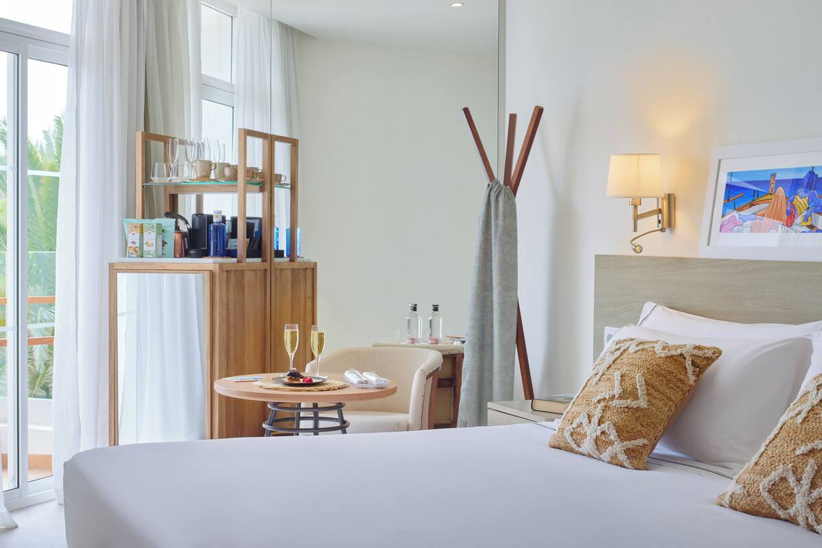 Ein Hotelzimmer mit einem Bett, einem kleinen runden Tisch mit einer Speisenplatte und zwei Weingläsern, einem Sessel, einem Kleiderschrank mit Regalen, einer Lampe und einem Gemälde an der Wand.