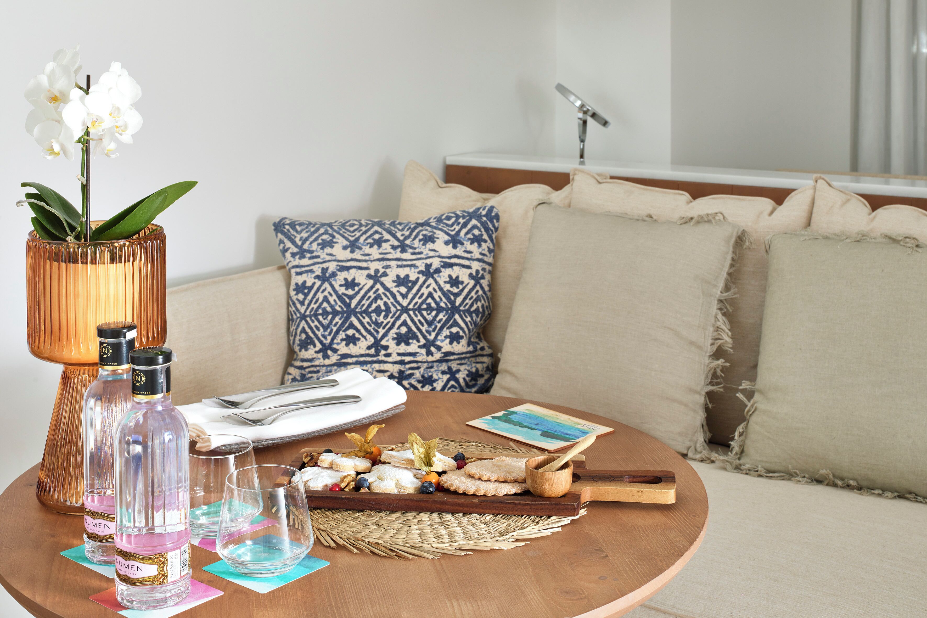 A round wooden table with a bottle of water, a glass, a napkin, a small wooden board with food, and a decorative vase with flowers.