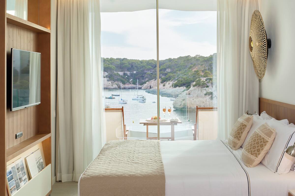 Ein Hotelzimmer mit einem großen Bett, einem Fernseher, einem Balkon mit Blick auf Boote und eine Klippe.