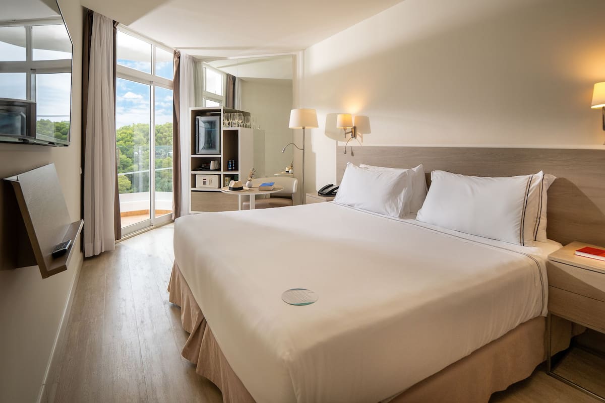 Ein Hotelzimmer mit einem großen Bett, Nachttischlampen, einem Schreibtisch mit Stuhl, einem an der Wand befestigten Fernseher und Blick ins Freie.