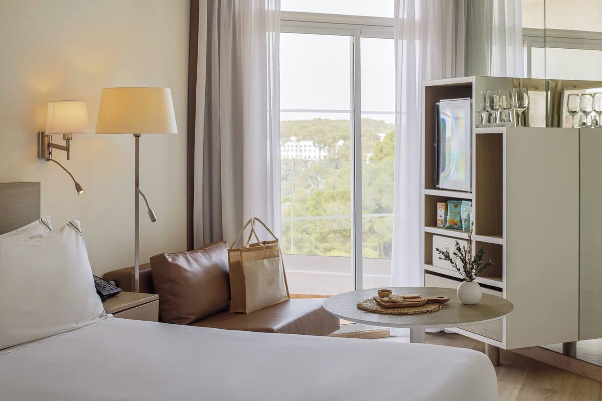 Ein Hotelzimmer mit Bett, Sofa, Nachttisch, Lampe, Tisch mit Vase und Blick auf Grün durch das Fenster.