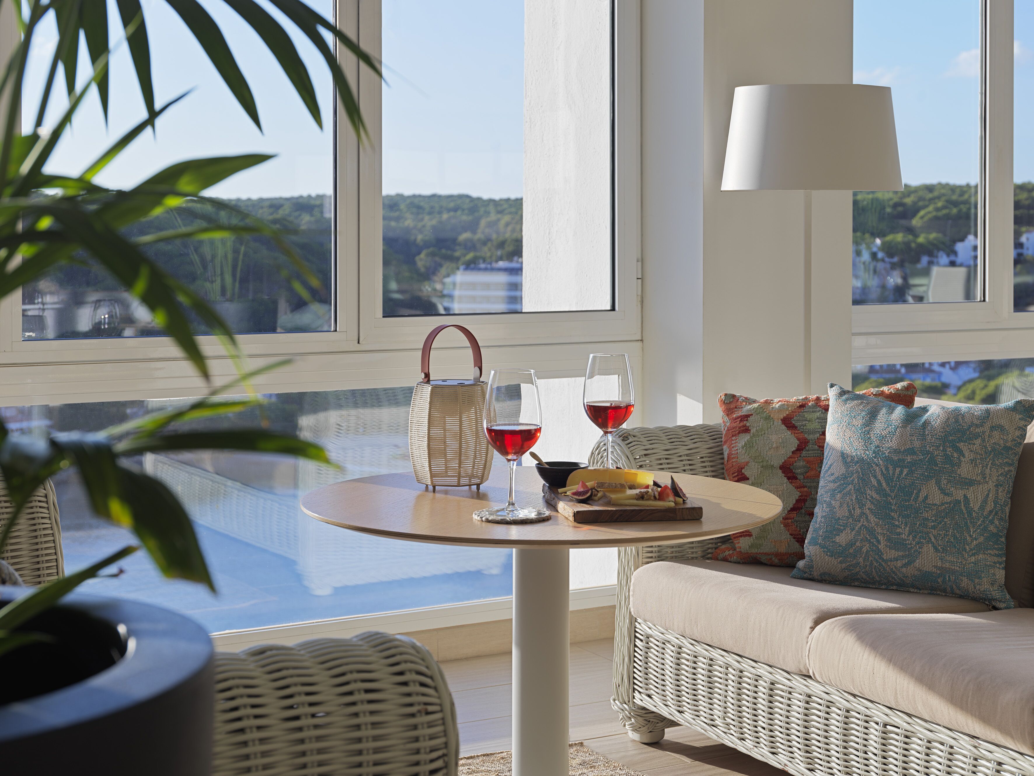 A sunlit room with a view of a lake, a wicker sofa, a table with wine glasses and a basket, and a lamp.
