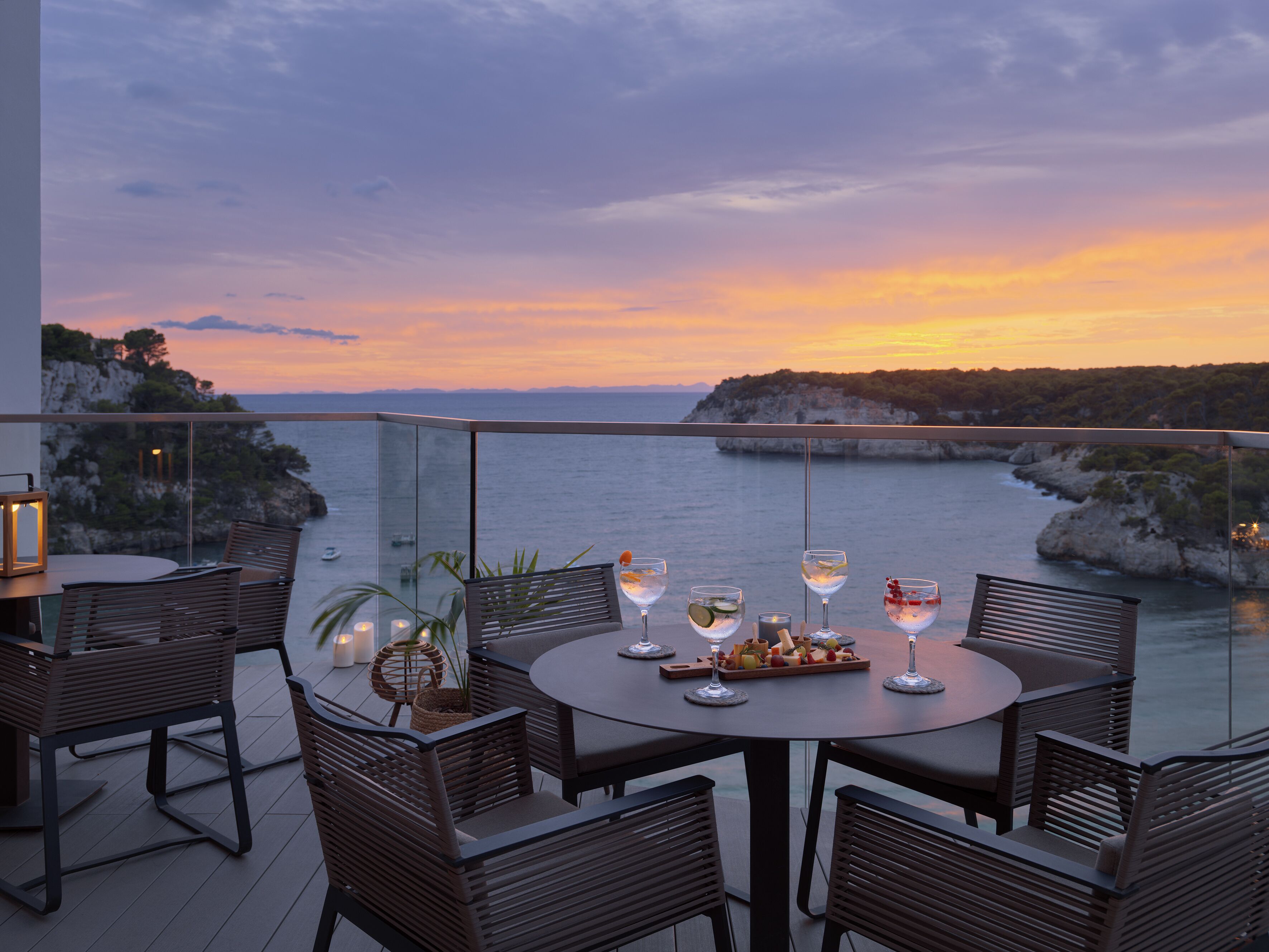 A round table with chairs and a glass railing overlooking a coastal view at sunset.