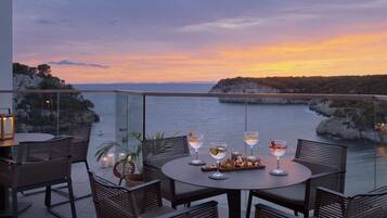 A round table with chairs and a glass railing overlooking a coastal view at sunset.