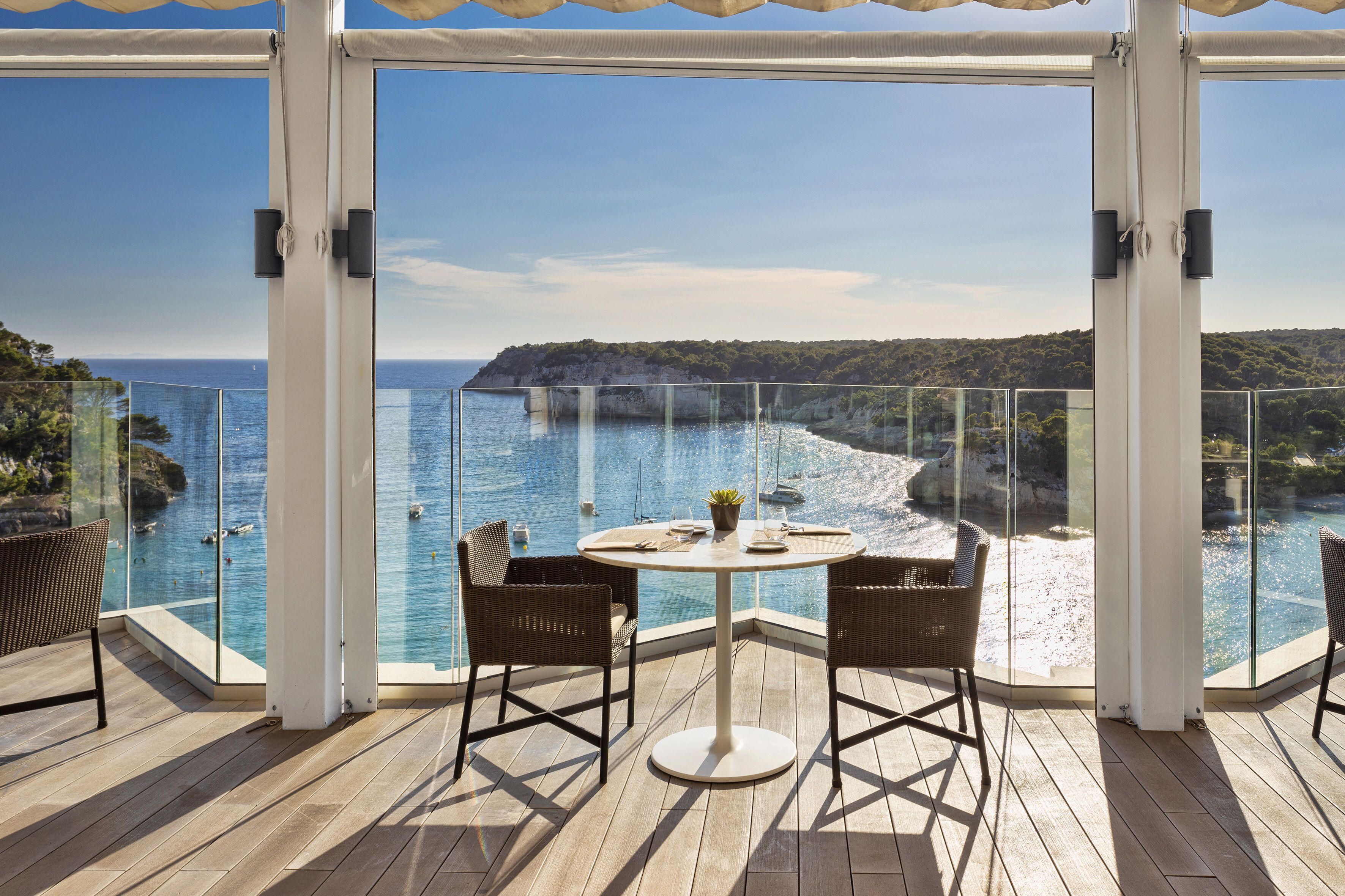 Uma varanda com guarda-corpo de vidro, uma mesa redonda com cadeiras e vista para o mar e falésias.