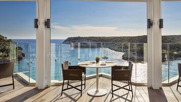 Uma varanda com guarda-corpo de vidro, uma mesa redonda com cadeiras e vista para o mar e falésias.