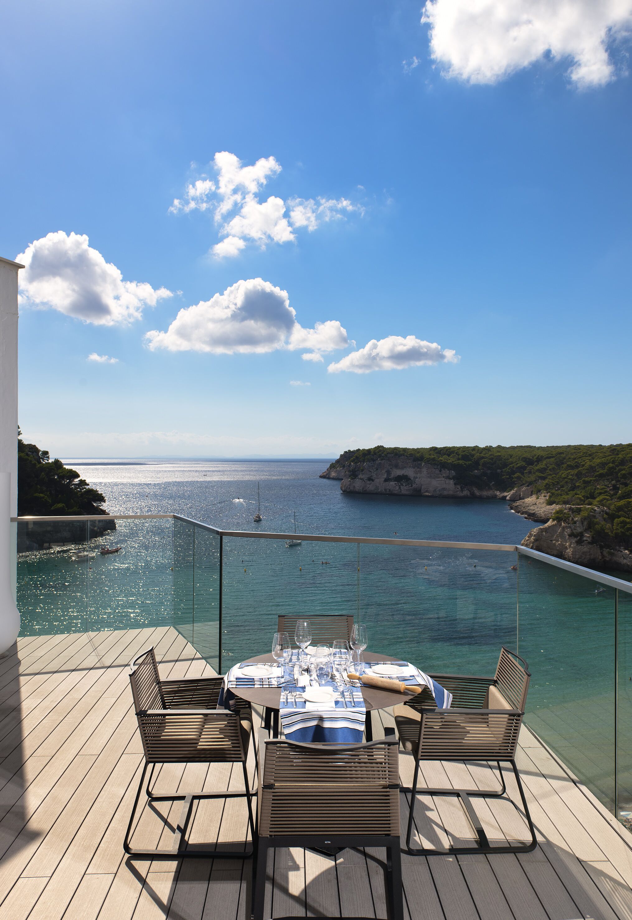 Un balcón con una mesa dispuesta para dos personas, con vistas a la costa, agua azul cristalina y acantilados distantes.
