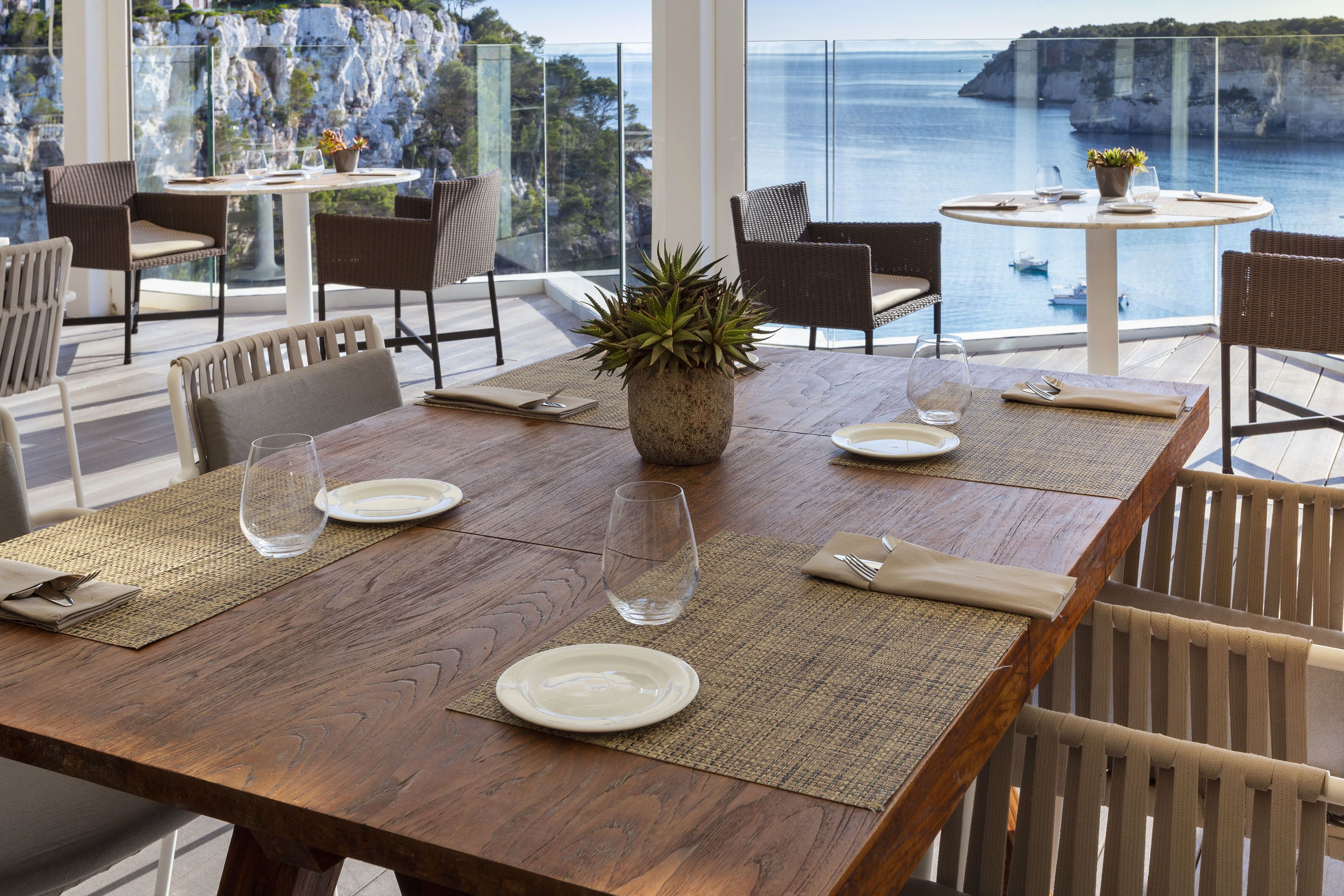 Comedor con una mesa de madera dispuesta para dos personas, rodeada de sillas de mimbre, con vistas al mar y a los acantilados.
